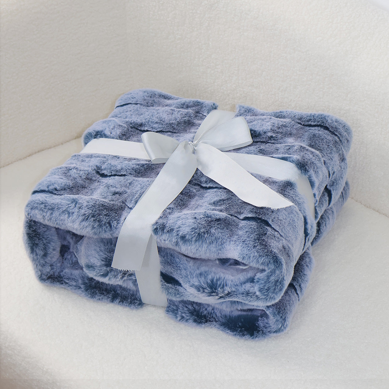 A folded soft blue plush blanket with a velvety texture, secured by a silver-gray satin ribbon tied into an elegant bow at the center. The clean light background creates a gentle contrast, emphasizing the blanket's cozy quality and refined presentation.