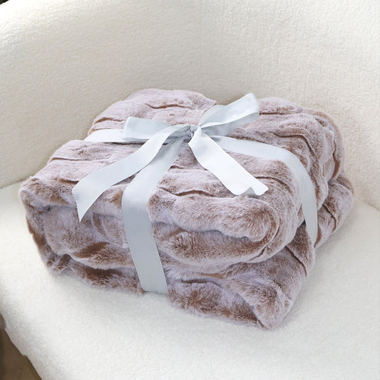 A stack of soft light brown plush blankets with a velvety texture, elegantly tied with a silver-gray satin ribbon bow at the top. The clean beige fabric background complements the blanket's warm tone, creating a harmonious and cozy aesthetic suitable for home decor display.