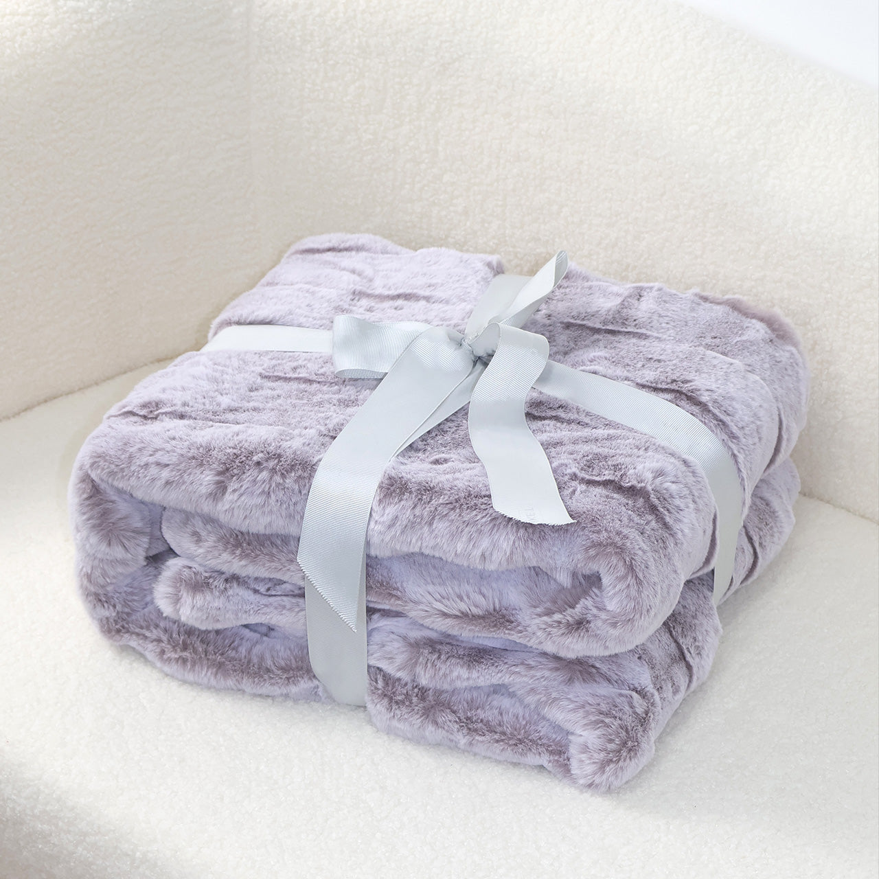 A folded lavender-gray plush blanket with a velvety texture, elegantly tied with a silver-gray satin ribbon bow. The soft off-white background creates a harmonious contrast, emphasizing the blanket's cozy appeal and sophisticated color palette.