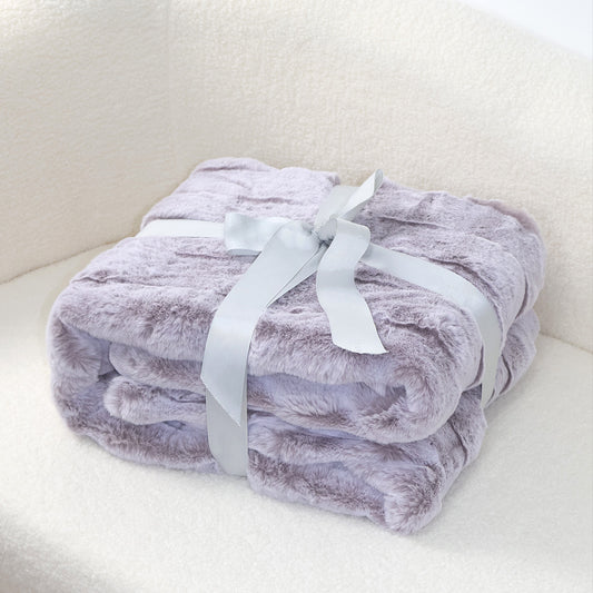 A folded lavender-gray plush blanket with a velvety texture, elegantly tied with a silver-gray satin ribbon bow. The soft off-white background creates a harmonious contrast, emphasizing the blanket's cozy appeal and sophisticated color palette.