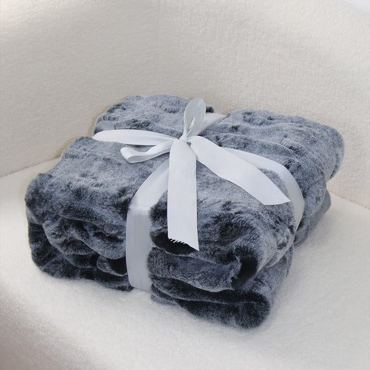 A rolled gray plush blanket with a soft, fluffy texture, secured with a white satin ribbon tied into an elegant bow at the top. The clean light background creates a subtle contrast with the blanket's neutral tone, emphasizing its cozy quality and refined presentation.