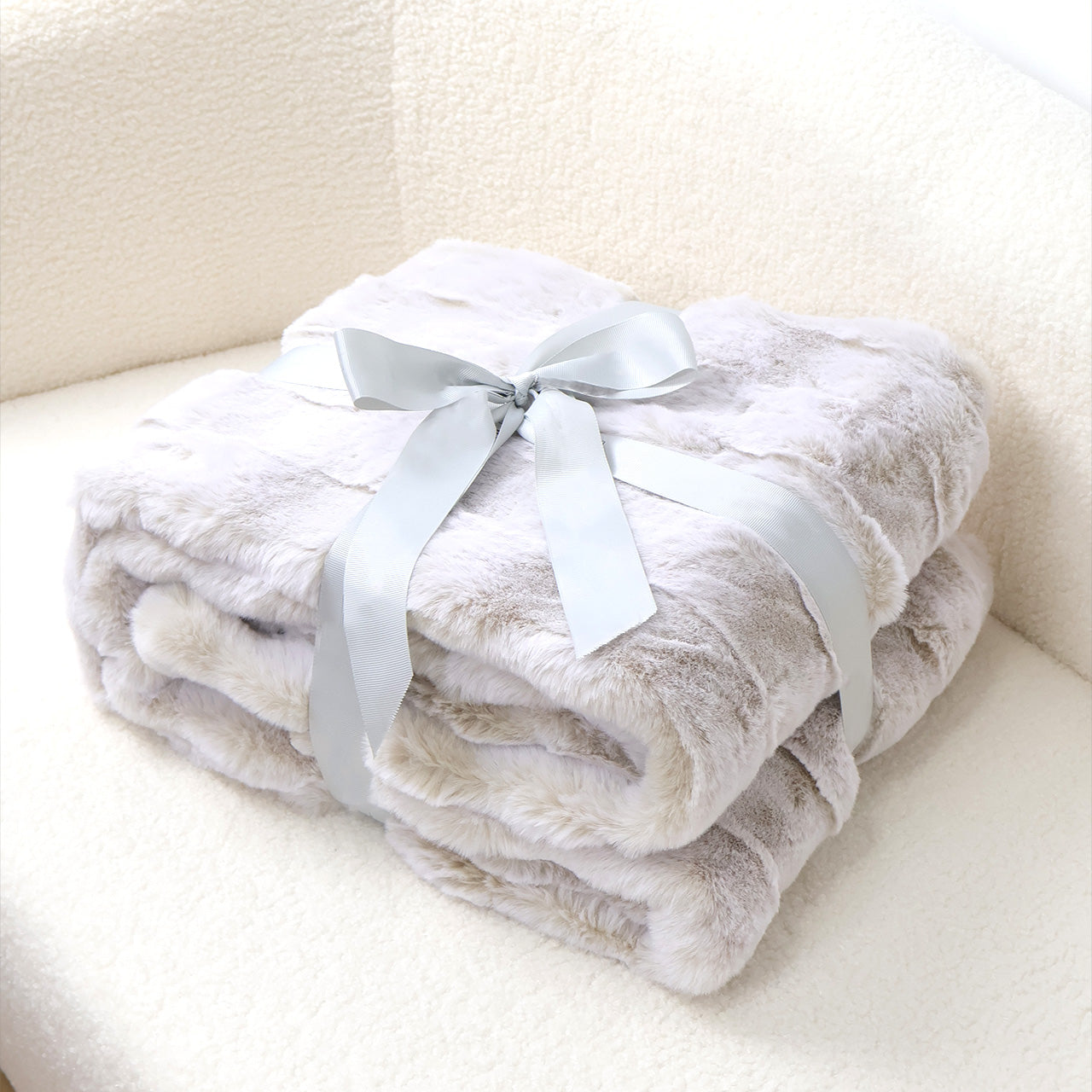 A stack of soft gradient-gray plush blankets tied with a silver-gray satin ribbon bow. The velvety texture and light gray-to-white color transition create a cozy aesthetic, complemented by a soft fabric background. The minimalist composition emphasizes the blankets' luxurious feel and versatile home decor appeal.