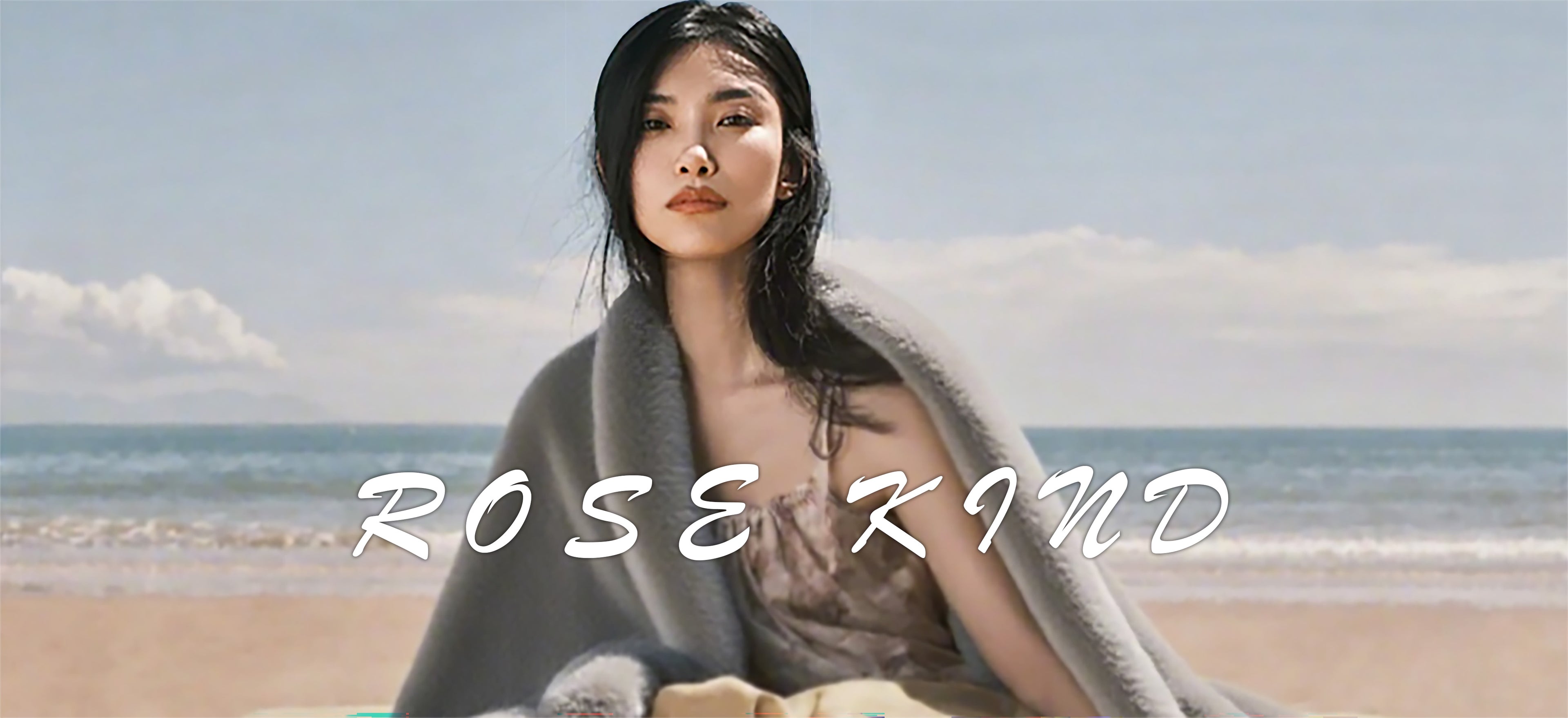 The image depicts a serene beach scene featuring a woman draped in a gray blanket, seated centrally with a muted color palette of sandy beaches and azure sea-sky backdrop. The brand identifier "ROSE KIND" appears in the foreground's lower-central position. Subtle visual depth is created through the blanket's flowing folds and the figure's silhouette, while gentle waves lapping the shore contrast with the subject's tranquil posture, evoking a calming elegance through its composition.