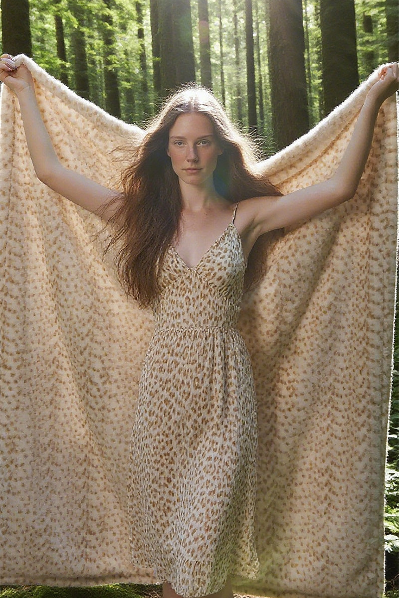 Female model in faux-leopard print V-neck sundress holding flowing fabric against forest backdrop. Dress features soft caramel spots on ivory base, with plush texture suggesting velour material. Sunlight filters through trees, creating dappled lighting on the natural ground.