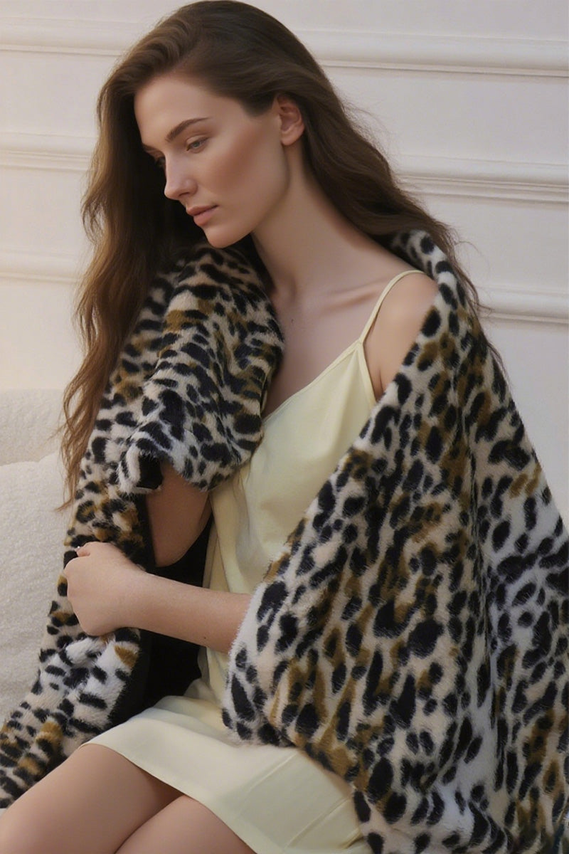 Female figure draped in faux-leopard print shawl with irregular caramel spots on ivory base. Shawl features soft velvet texture and flows asymmetrically over a black top. Background suggests indoor setting with neutral tones, emphasizing the garment's luxurious drape.