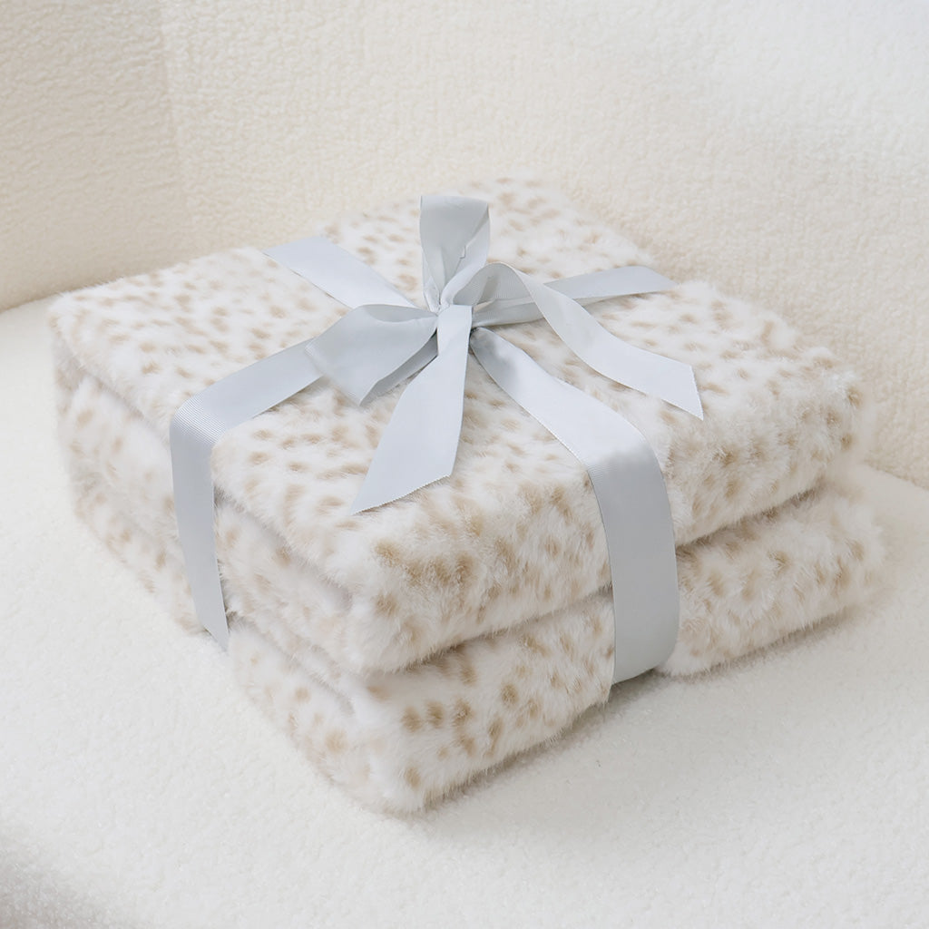 Double-layered faux-leopard print blanket stack in ivory with soft caramel spots, featuring plush velvet texture and silver-gray satin bow accent. Neatly arranged on light-toned furniture surface, suggesting luxury home decor or thoughtful gift presentation.