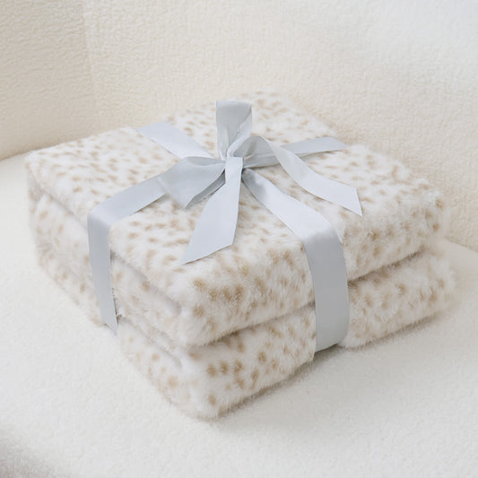 Double-layered faux-leopard print blanket stack in ivory with soft caramel spots, featuring plush velvet texture and silver-gray satin bow accent. Neatly arranged on light-toned furniture surface, suggesting luxury home decor or thoughtful gift presentation.