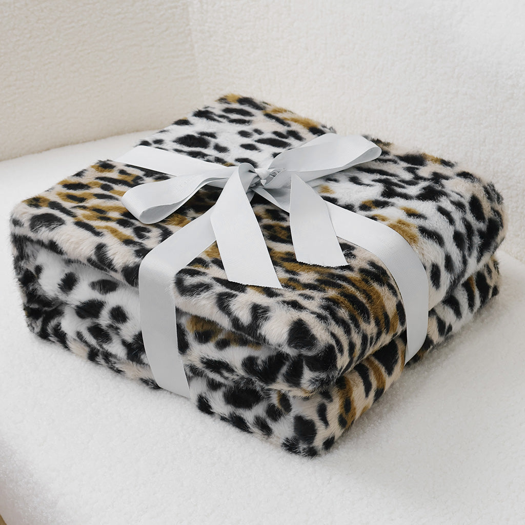 Neatly folded faux-leopard print blanket in black, brown, and ivory tones, featuring plush velvet texture with white satin ribbon bow. Centered on light neutral background, suggesting versatile use as home decor or cozy layering piece.