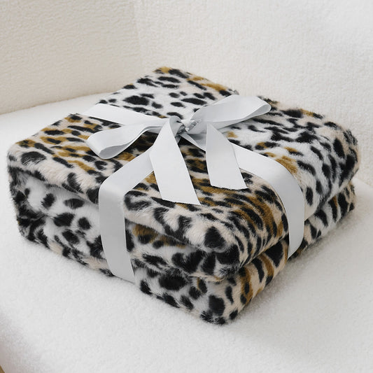 Neatly folded faux-leopard print blanket in black, brown, and ivory tones, featuring plush velvet texture with white satin ribbon bow. Centered on light neutral background, suggesting versatile use as home decor or cozy layering piece.