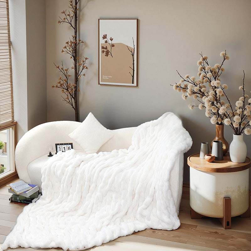 A white textured blanket drapes over a curved white sofa with a cushion. Background includes a side table with vases, artwork, and a window with blinds.