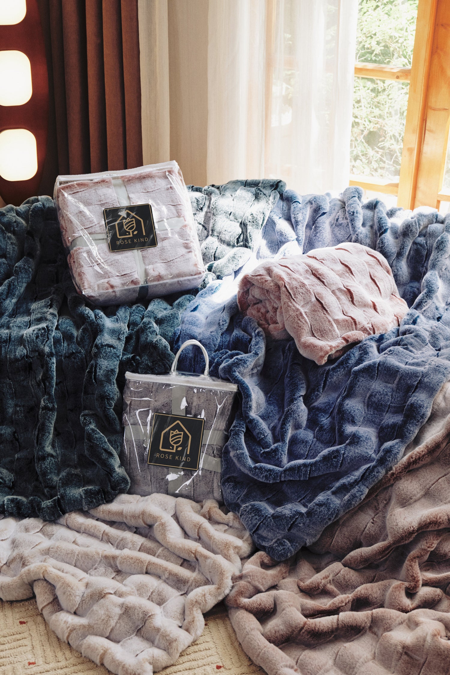 A cozy indoor scene with knit throws in navy, blue, pink, gray, and beige, casually draped and rolled. Soft light from a window with greenery outside. "ROSE KIND" tags visible. Warm, relaxed vibe.