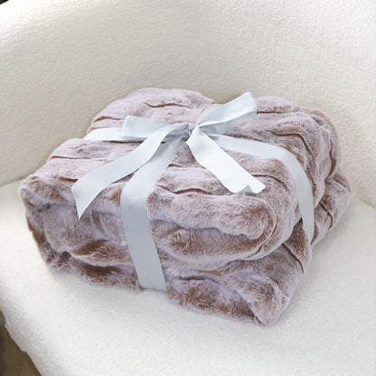 A stack of soft light brown plush blankets with a velvety texture, elegantly tied with a silver-gray satin ribbon bow at the top. The clean beige fabric background complements the blanket's warm tone, creating a harmonious and cozy aesthetic suitable for home decor display.